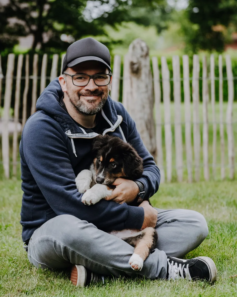 Maik Haring mit Hundewelpe im Garten, lächelnd, mit Cap und Brille